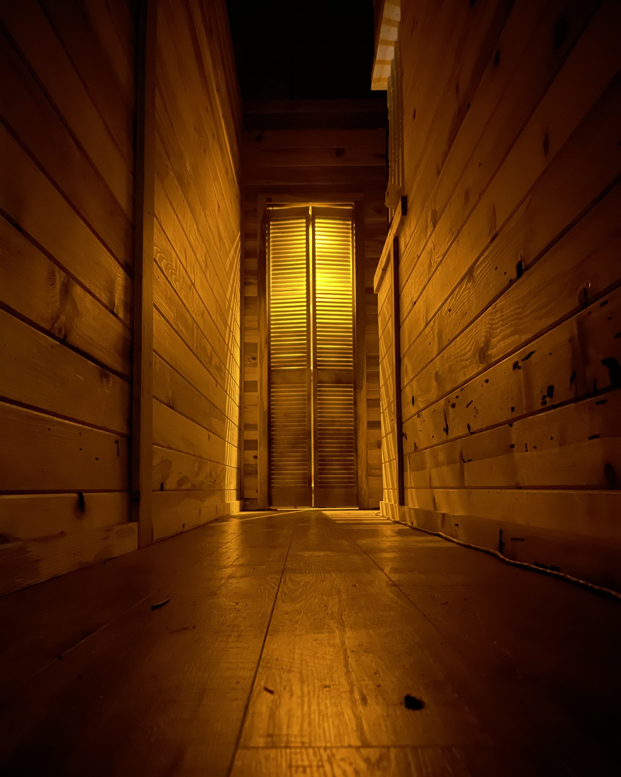 Narrow wooden corridor in an escape room set leading to a lit louvred door